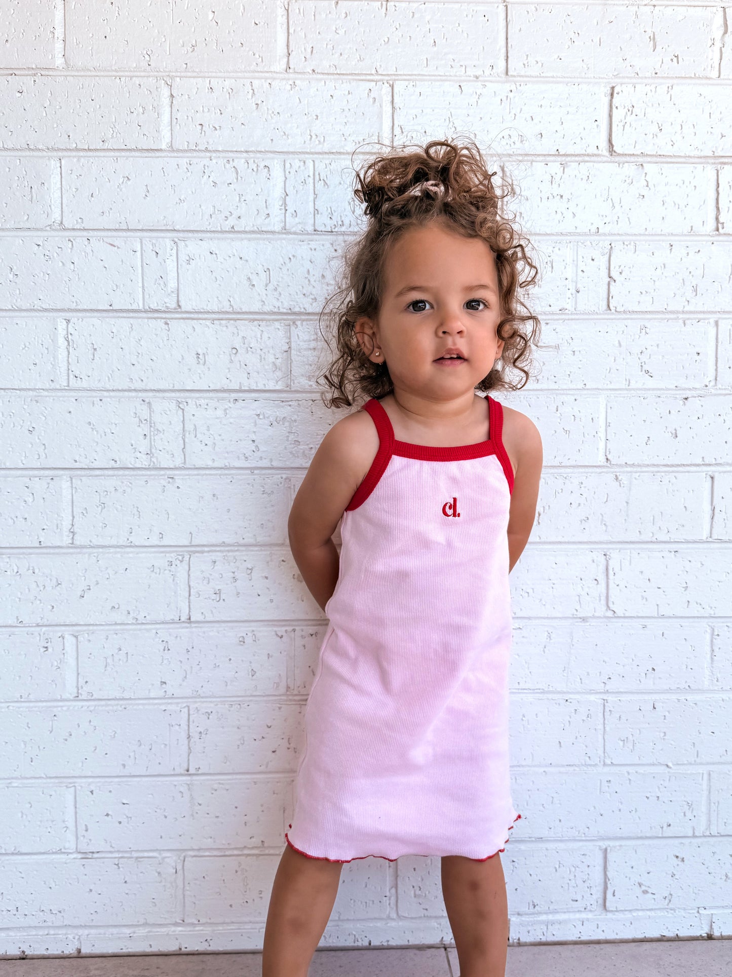 Child wearing a pink dress with red straps (Cami Lane Essential Ribbed Dress) against a white brick wall.