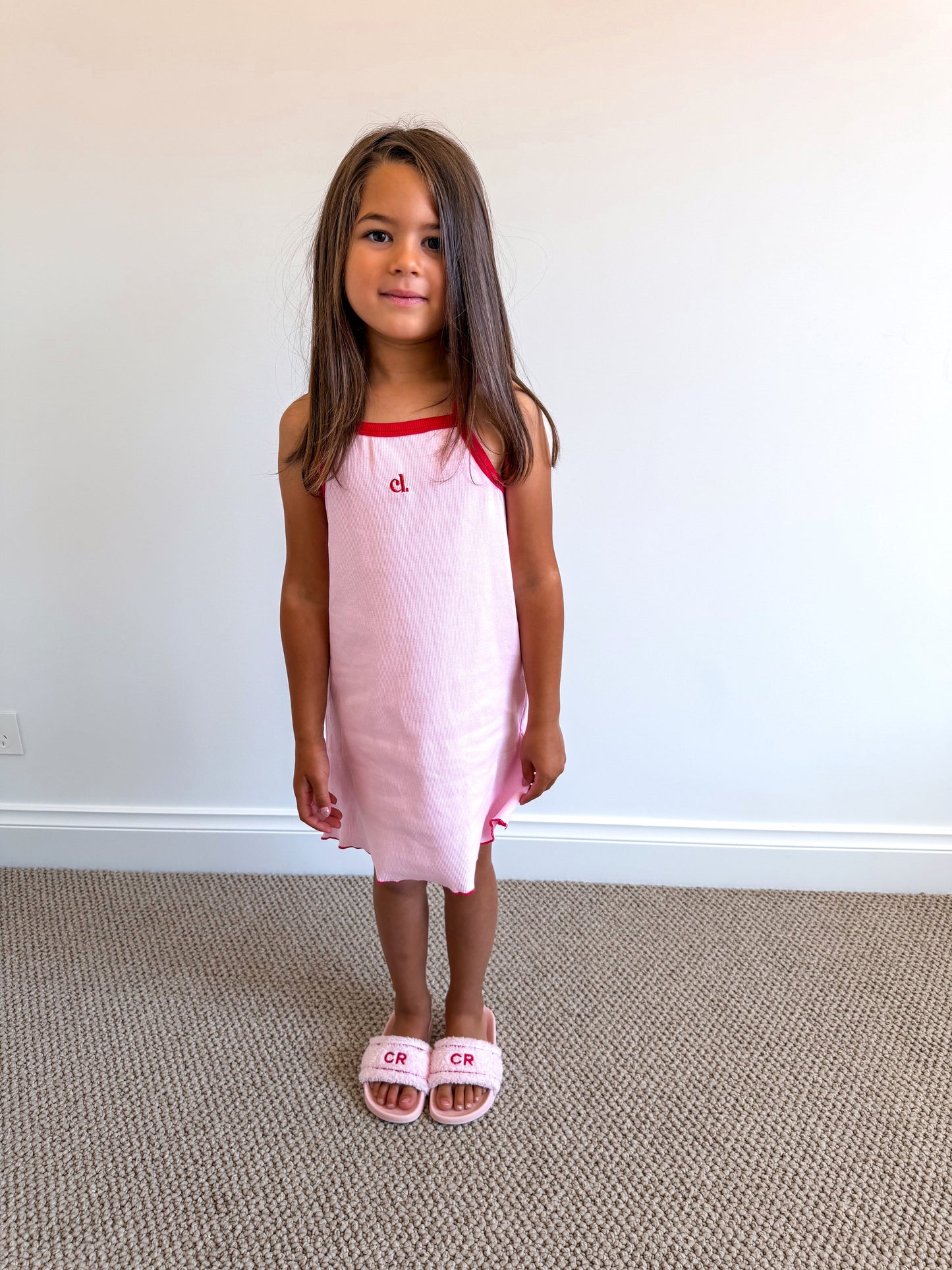 Young girl wearing a pink dress with a red neckline (Cami Lane Essential Ribbed Dress) against a plain background.