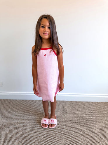Young girl wearing a pink dress with a red neckline (Cami Lane Essential Ribbed Dress) against a plain background.