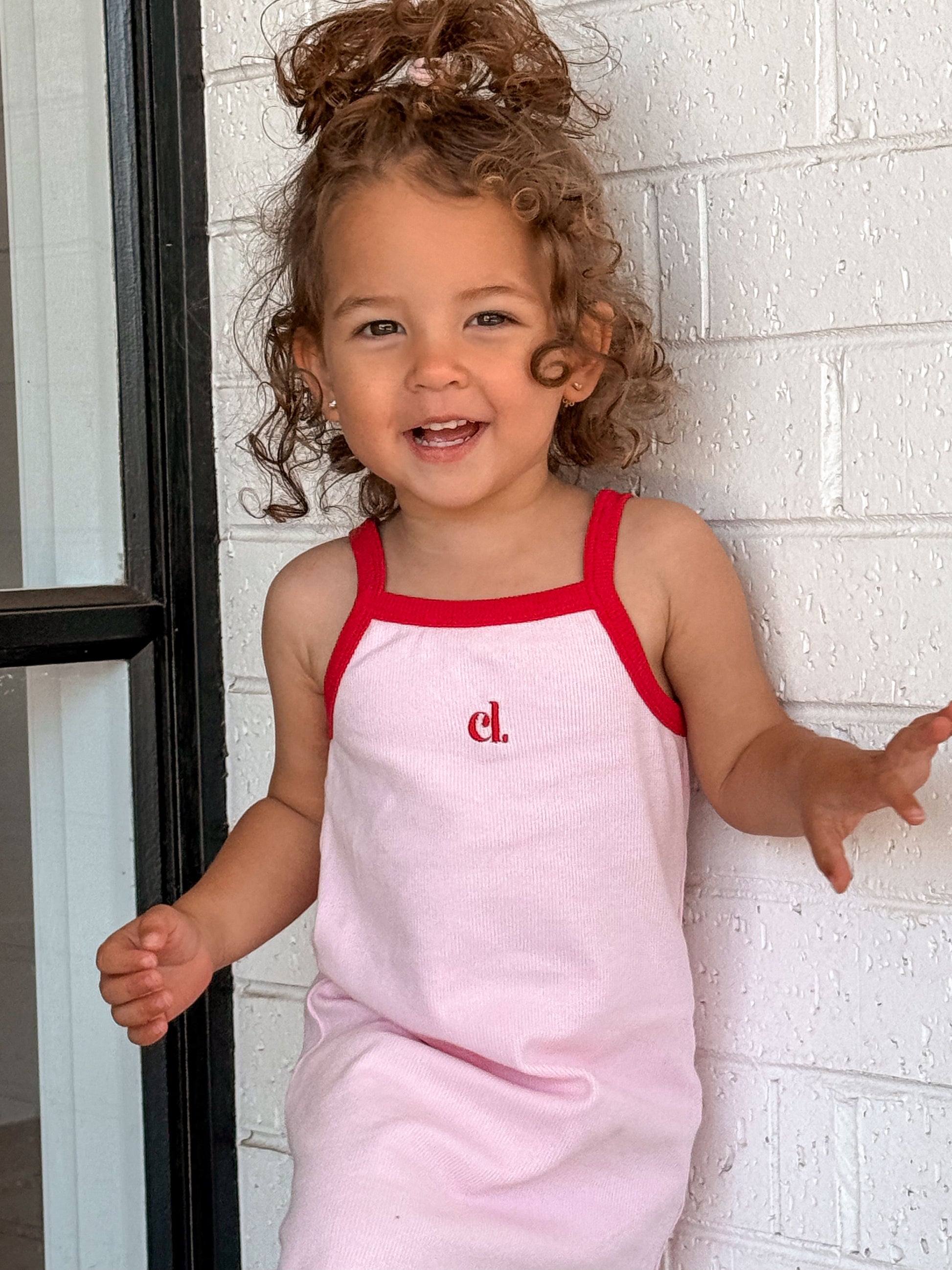 Child wearing a pink dress with red straps (Cami Lane Essential Ribbed Dress) against a white brick wall.