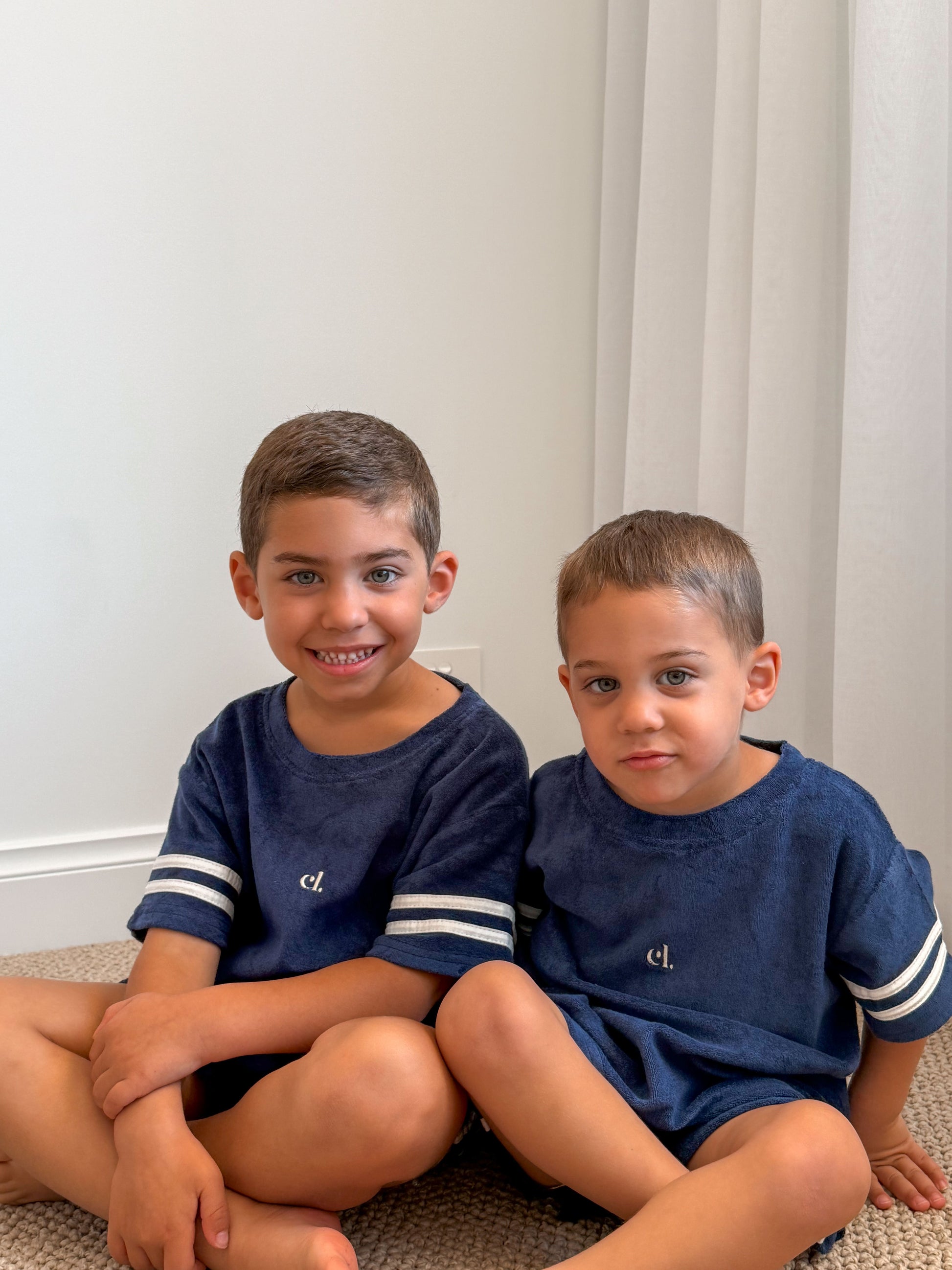 Two young boys in matching navy Cami Lane Soft Terry Set outfits sitting down against a white wall.