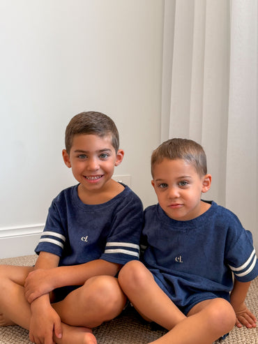 Two young boys in matching navy Cami Lane Soft Terry Set outfits sitting down against a white wall.